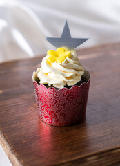 Cupcake with piped vanilla frosting, yellow flower decoration and paper star topper, placed on wooden board in clean setting, macro dessert photo with soft light for menu visuals, bakery branding