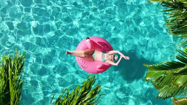 A vibrant aerial scene featuring a woman floating on a pink flamingo float in a crystal-clear blue pool, surrounded by palm trees, bright loungers with towels and striped umbrella on a warm sunny day