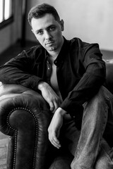 Obraz premium Frontal portrait of young self confident handsome young man with naked torso in a black shirt in a stylish loft interior, grey walls