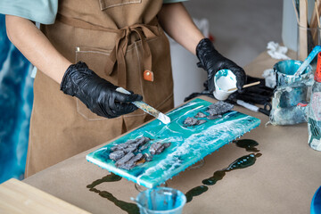 Craftswoman artist hands in gloves use wooden spatula to create waves in epoxy resin in workshop....