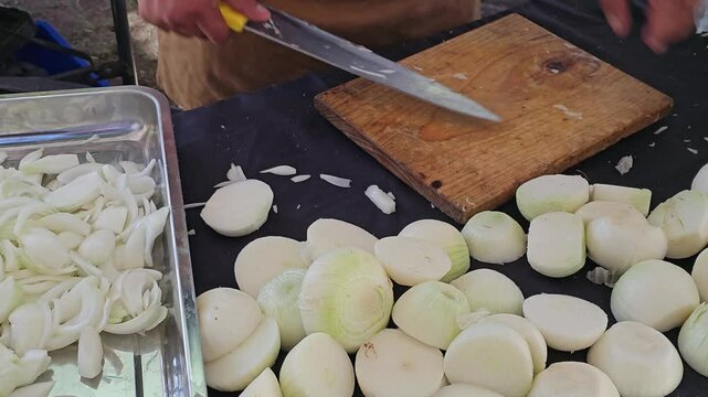 A cook at a Chapala food stall chopping fresh onions on a wooden cutting board. Large stacks of tortillas and trays of sliced onions are visible, capturing the preparation for a local culinary event.