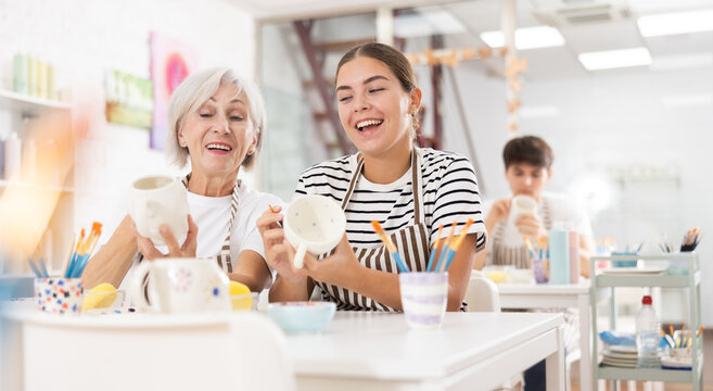 Cheerful young girl working together with elderly mother in family pottery studio, chatting and laughing carefree while creating unique paint designs on ceramic cups.. - Powered by Adobe