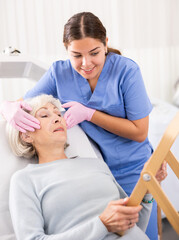 Positive aged woman patient lying on clinical chair discussing face aesthetic procedures with a...