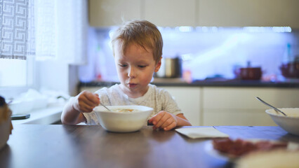 Blond toddler eating hot soup from a white bowl, sitting at the kitchen table in a bright modern...