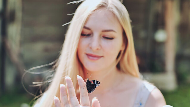 Blonde woman gently holding colorful butterfly on fingertip, experiencing tranquil garden moment under bright sunlight