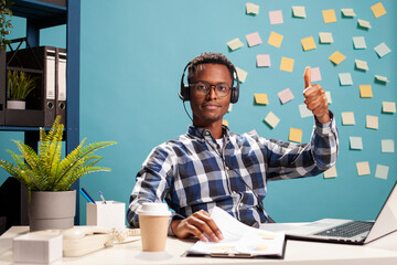 Customer support agent wearing a headset and giving a thumbs up, seated at desk reviewing...