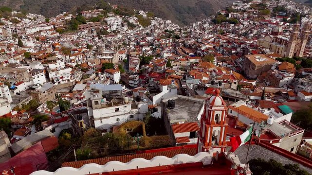 Panoramic view of Taxco de Alarcon in Mexico, with cathedral in background and church with Mexican flag in foreground. Stationary drone camera shot