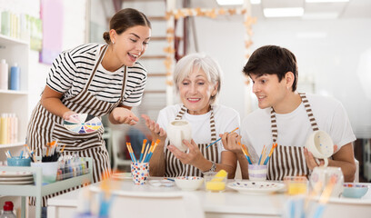 Cheerful elderly woman, along with young girl and guy wearing aprons engaging in ceramic dishware...