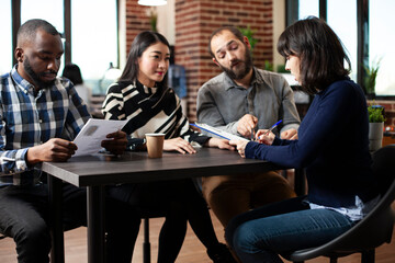 White woman signing her contract on clipboard with assistance from diverse human resources team in brick wall office. Female candidate completing her employment application during interview meeting.