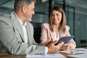 Smiling older professional executive business man talking working in corporate office at meeting with female bank manager consulting client looking at digital tablet computer discussing sales project.