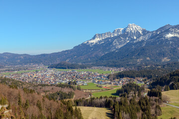 Naklejka premium A view of Reutte from the Highline 179 suspension bridge in Tyrol, Austria, reveals snow-capped valleys, dense forests, and towering peaks beneath a clear sky, offering a panorama of nature’s beauty.