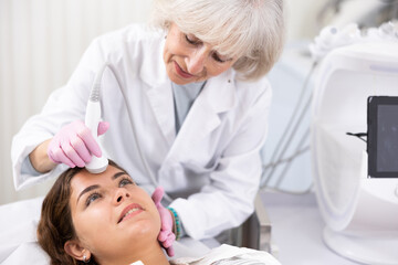 Positive young woman having face laser therapy by means of apparatus for aesthetic procedures used by specialist