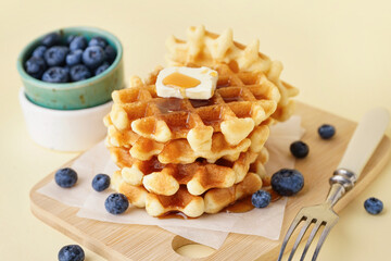 Wooden board of tasty waffles with blueberries and maple syrup on yellow background