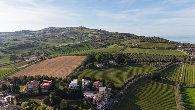 An aerial view showcases a vibrant Italian landscape featuring rolling green hills, vineyards, scattered houses, and a distant coastline under a clear sky.