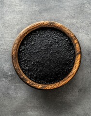 Black powder in a wooden bowl on a grey surface, top-down view. High-resolution studio photography with sharp focus and detailed texture.