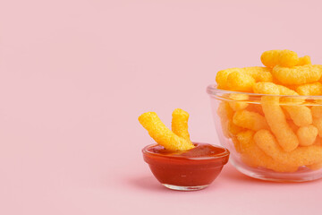 Glass bowl with tasty salty corn sticks and ketchup on pink background, closeup