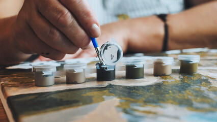 Artist's hand hovering near paint pot, grasping paintbrush, preparing to fill numbered canvas section with vibrant color during creative hobby activity