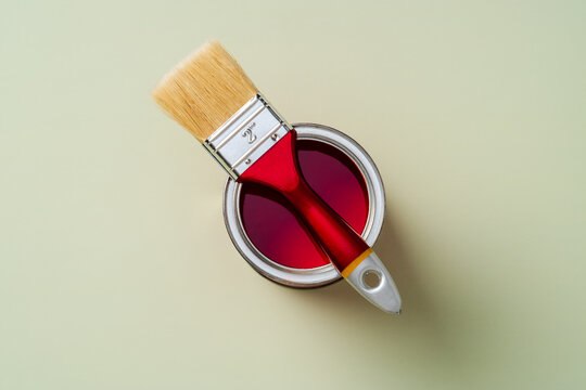 Top view of a red paintbrush resting on an open can of vibrant red paint against a light green background. Ideal for DIY, renovation, or art projects.