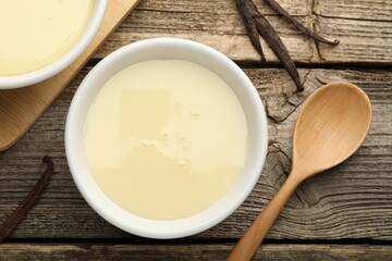 Tasty condensed milk, spoon and vanilla pods on wooden table, flat lay