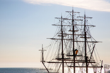 Silhouette of tall sailing ship with multiple masts and rigging against a calm sea at sunset. The sky glows with soft blue and orange hues, reflecting on the water, creating a historic maritime scene