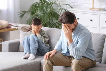Thoughtful young father and his sad little son sitting on sofa after squabbling at home