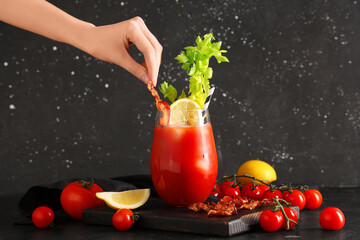 Female hand with bacon and glass of bloody mary cocktail with lemon on black background