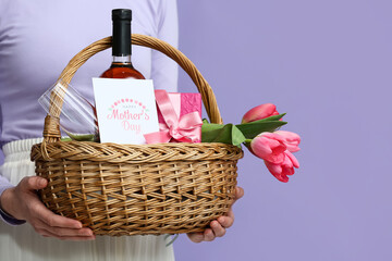 Young woman holding basket with wine bottle, tulips and gift box for Mother's Day on lilac...