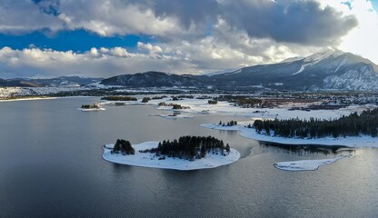 Aerial view of Dillon Reservoir in Frisco, Colorado, USA, showcasing snow-covered mountains, islands, and the town. The sun shines through the clouds, creating a scenic winter landscape.