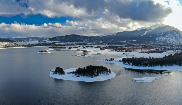 Aerial view of Dillon Reservoir in Frisco, Colorado, USA, showcasing snow-covered mountains, islands, and the town. The sun shines through the clouds, creating a scenic winter landscape.