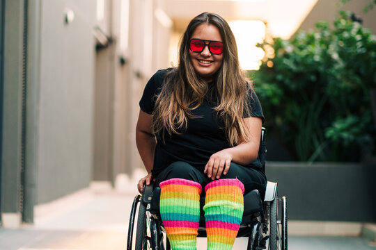 Beautiful Latina woman in a wheelchair wearing retro red sunglasses and multicolored tights, smiling and raising her arms in a gesture of joy. Wellness and health