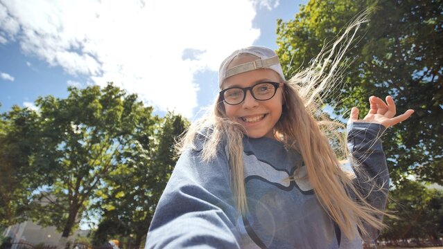 Blond haired schoolgirl wearing glasses and baseball cap, capturing selfie under bright sunlight with cheerful expression