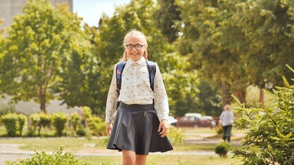 Cheerful schoolgirl with glasses walking through sunny park, backpack slung over shoulder