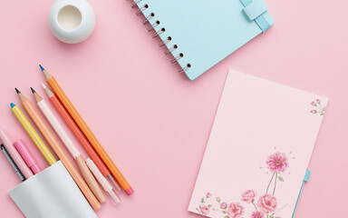 Pastel stationery items are arranged on a pink surface, including a spiral notebook, colored pencils in a holder, and a small white round object. Flat lay, top view.