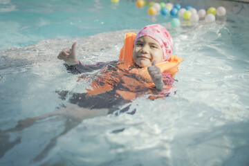 A young girl is in a pool wearing a life jacket and a pink hat. She is smiling and giving a thumbs...