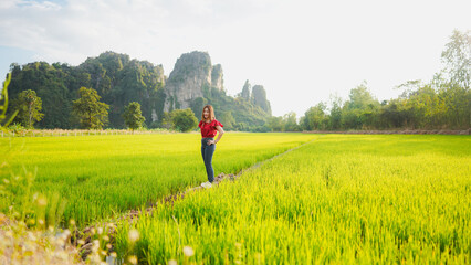A woman stands in a vibrant green rice field, surrounded by stunning mountains and a clear sky,...