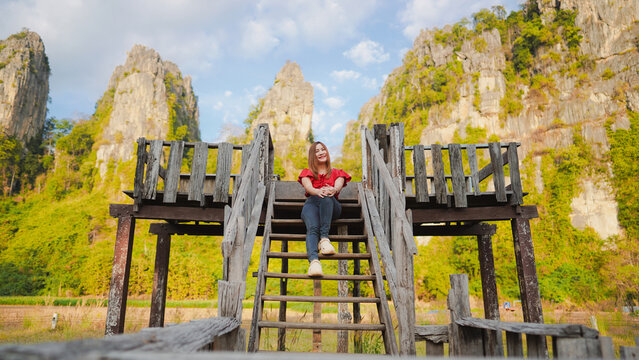 A serene landscape featuring a woman sitting on a wooden observation deck surrounded by towering rock formations and lush greenery.