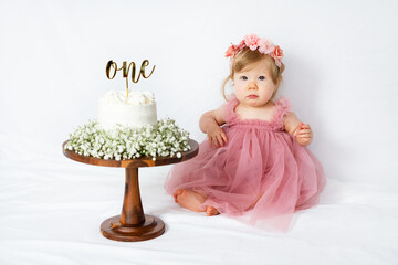 Portrait of an Adorable baby girl's first birthday cake smash in a pink tulle dress with a flower crown. Baby girl sits beside a white frosted cake topped with "one"