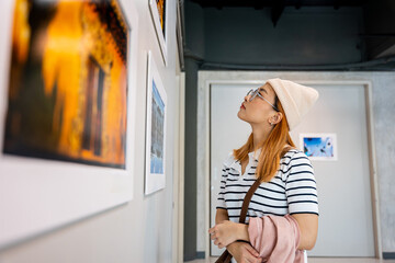 Asian woman standing she looking art gallery in front of colorful framed paintings pictures on white wall, people watch at photo frame to leaning against at show exhibition artwork gallery, Side view