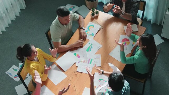 Top view of smart people throw paper and giving high five at meeting table with financial document and stock market analysis paper scattering around. Aerial view of team celebrate project. Symposium.