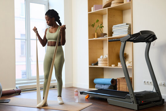 Woman performing resistance band exercises in modern home gym setting featuring treadmill and well-organized shelves filled with various items