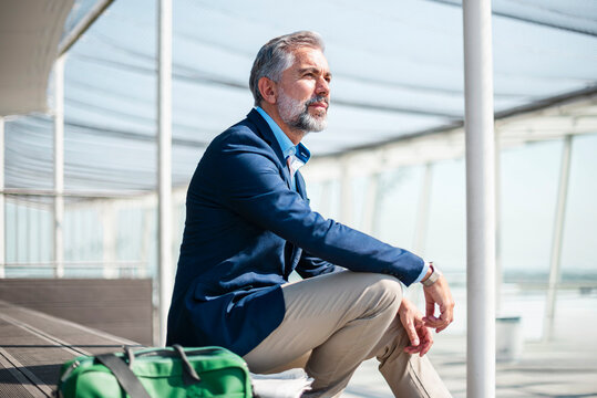 Mature businessman sitting outdoors with a travelling bag, looking thoughtful