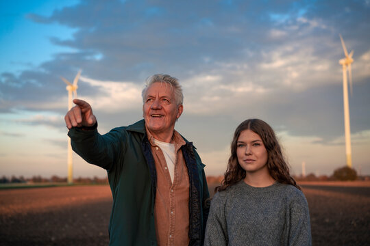 Grandfather and granddaughter in a field with wind turbines at sunset
