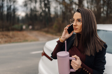 Businesswoman making a phone call while holding a travel mug near her car on the side of the road