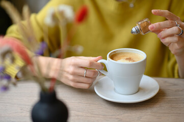 A woman’s hands sprinkling cinnamon into a cup of coffee at a cozy café. A warm, inviting scene capturing the essence of comfort, relaxation, and the simple pleasures of a coffee break.