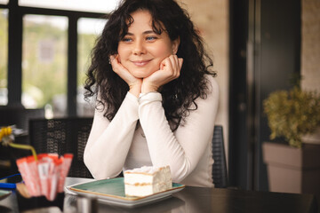 Happy woman sitting in cafe, hold her head and look at side. Attractive curly brunette dreaming girl rest in restaurant. Cream cake krempita on the table.
