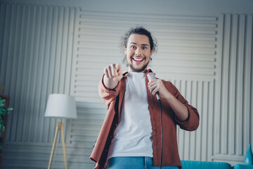 Happy young man enjoying karaoke at home in a cozy living room, holding a microphone and pointing...