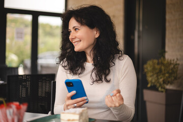 Joyful curly brunette woman playing video game on mobile phone, reading good news, celebrating...