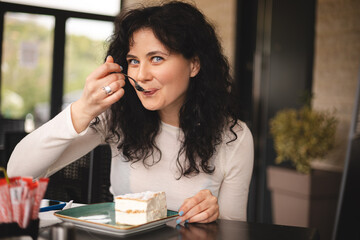 Young curly brunette woman eating cake with cream using fork sitting in cafe. Cheat meal day...