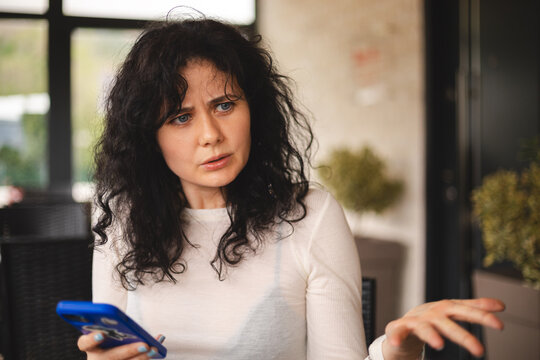 Beautiful upset angry curly woman sitting alone near big window in coffee shop, relaxing in restaurant during free time. Sad angry female read bad massage with mobile phone, yelling. Screaming woman.