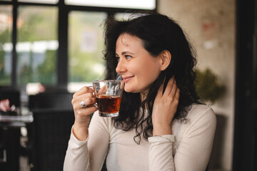 Happy brunette curly woman sitting in cafe and enjoy of smell and taste of tea, when have good conversation with someone. Woman in white long drink tea, she is smiling. 